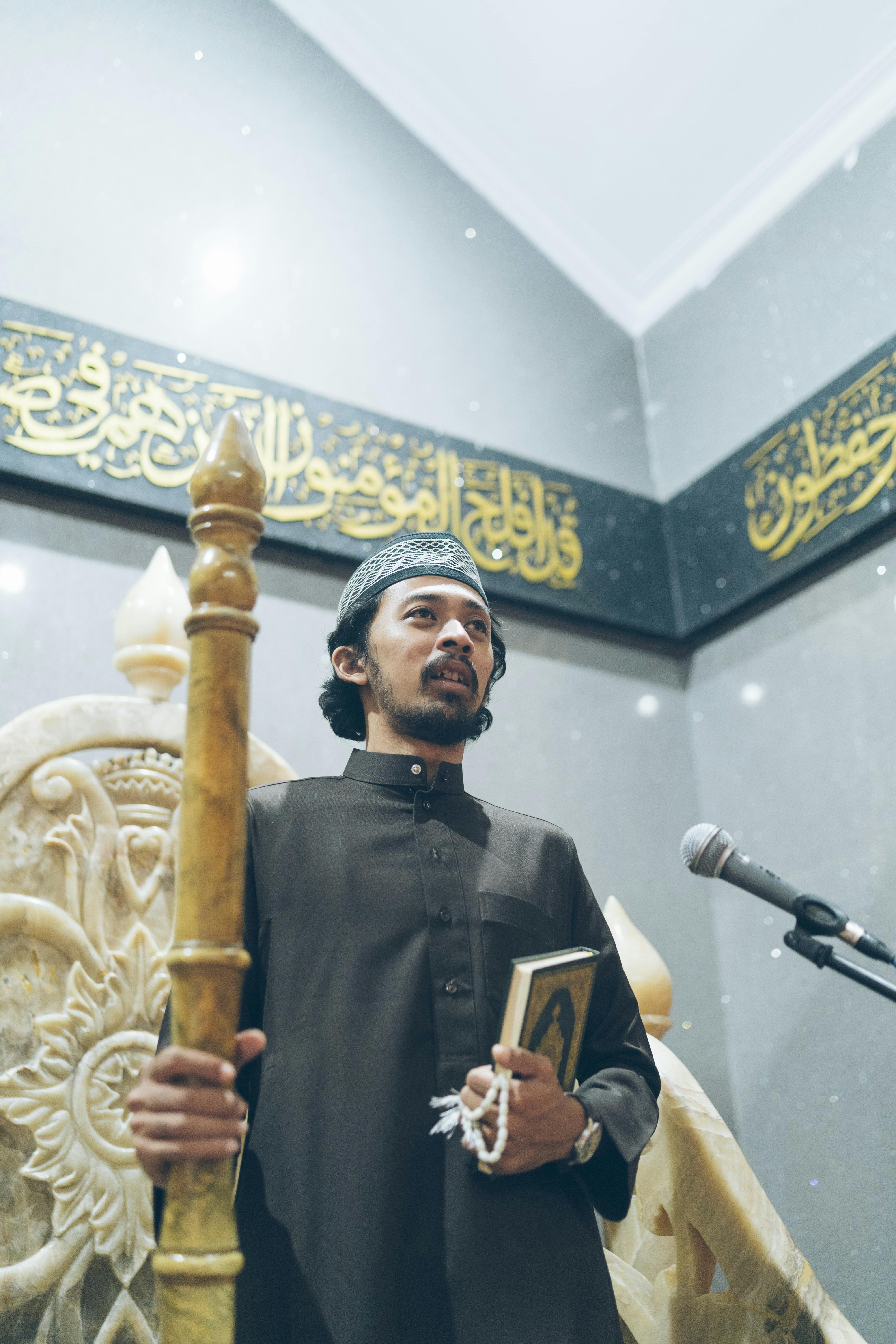 A man in traditional attire holding the Quran inside a mosque, standing near a microphone.