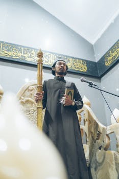 A religious leader gives a sermon in a mosque, holding a Quran, surrounded by intricate marble decor.