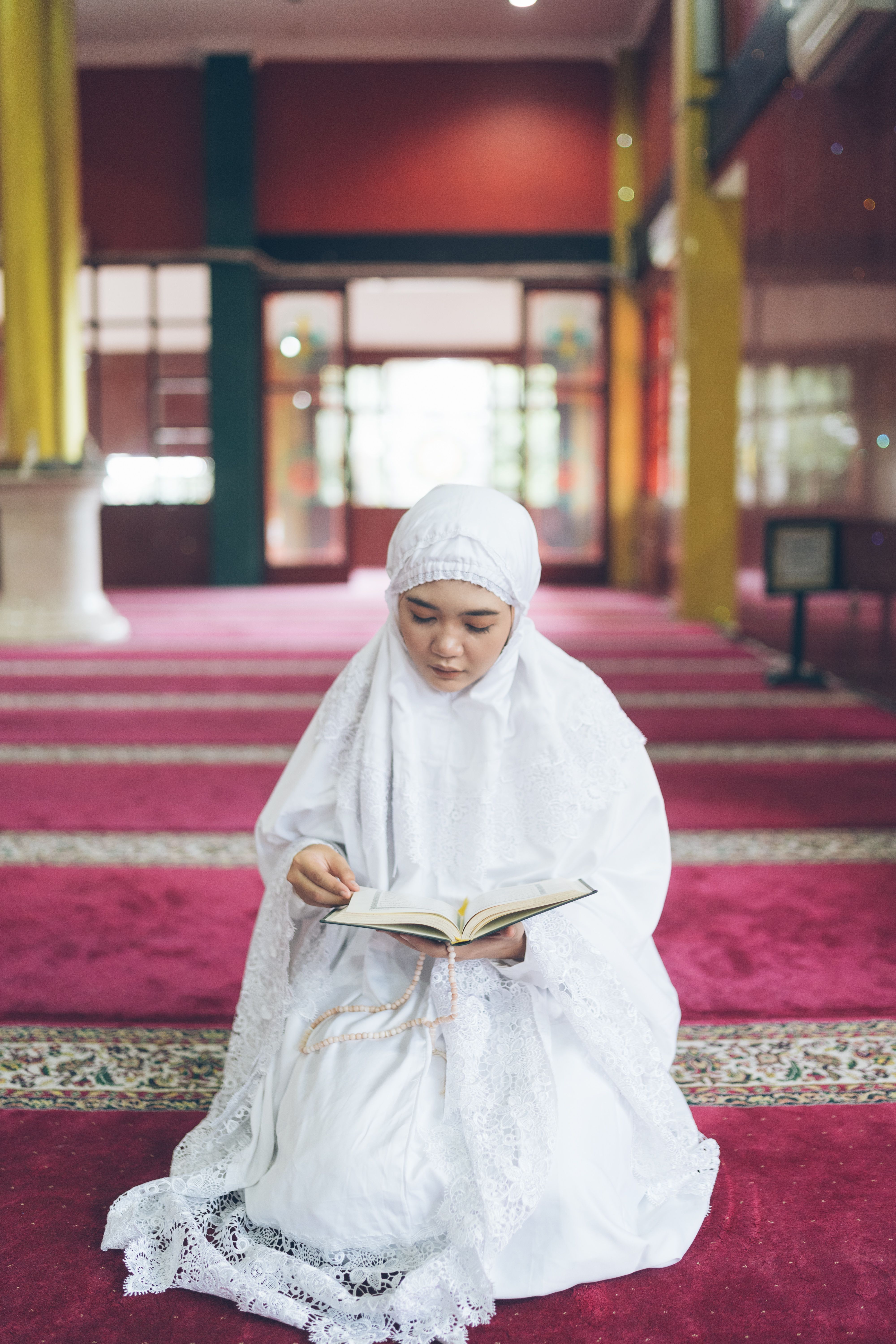 Young woman in white hijab reading the Quran inside a mosque, symbolizing spirituality and devotion.