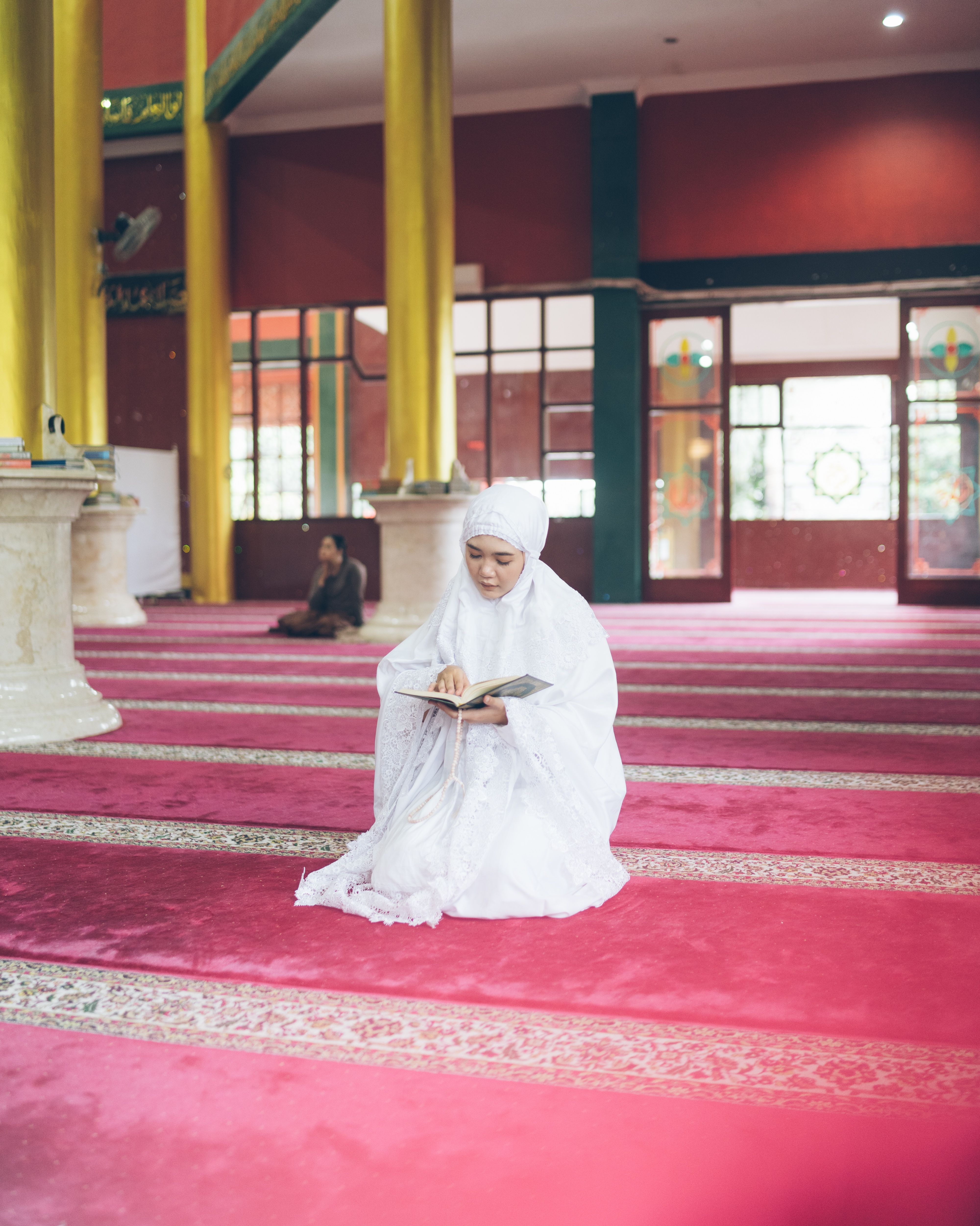 Woman wearing white hijab reads Quran in a mosque, highlighting spiritual devotion.