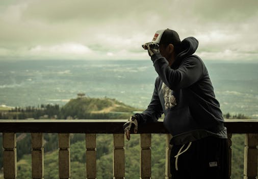 A man in casual attire leans on a railing, enjoying a scenic mountain view.