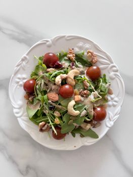 Fresh arugula salad topped with assorted nuts, cherry tomatoes, and creamy dressing on a white plate.