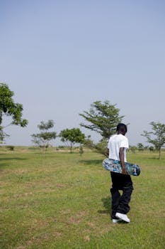 A young man carrying a skateboard strolls through a green park on a sunny day.
