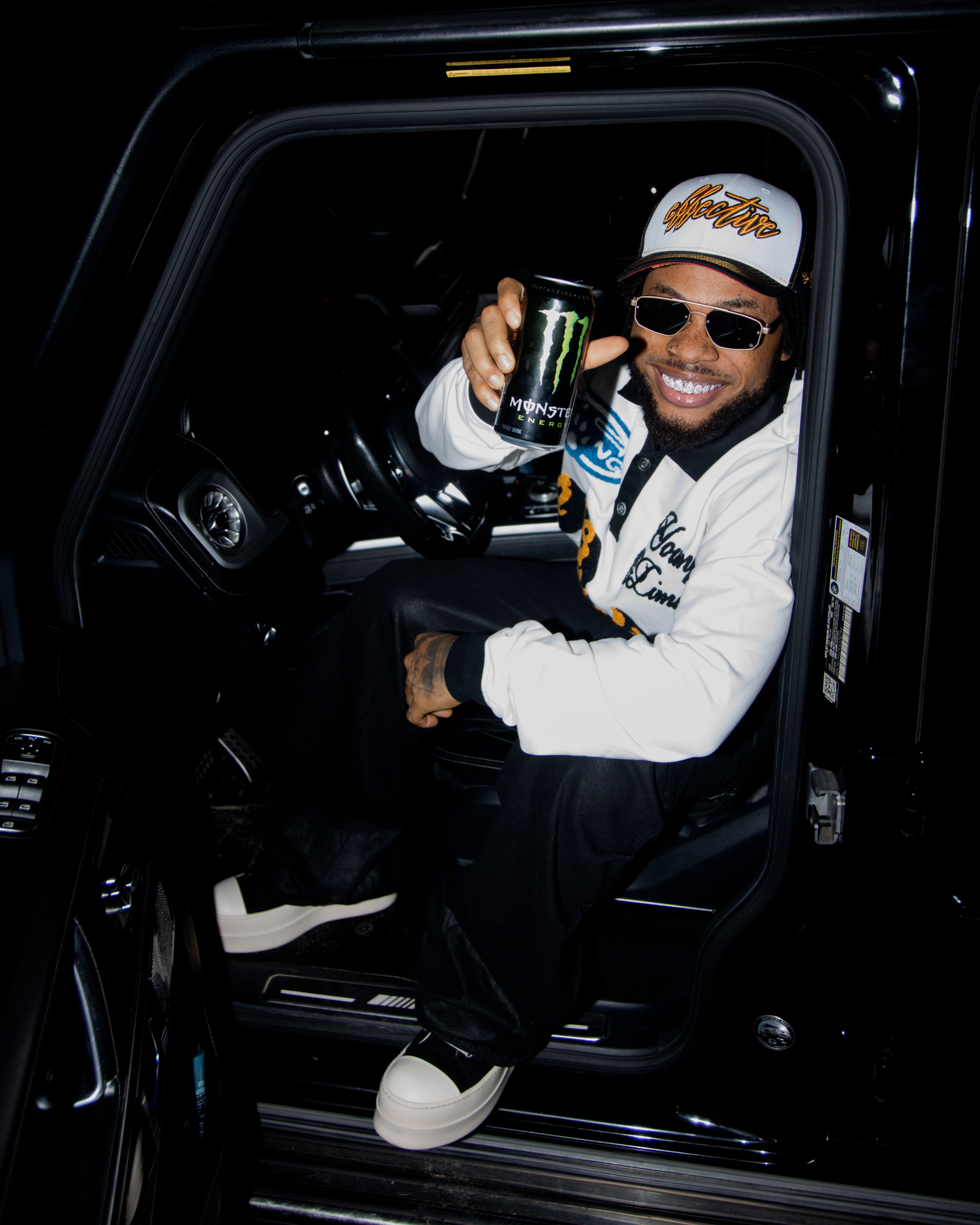 Smiling man in car holding Monster Energy drink, wearing sunglasses and cap at night.