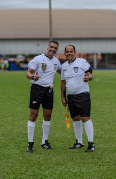 Two smiling soccer referees giving thumbs up on a grassy field.
