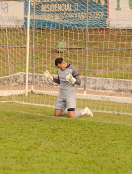 Soccer goalkeeper in gray jersey kneeling in front of goal net during a daytime match.