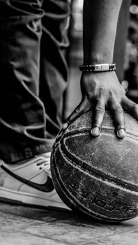 Monochrome image of a basketball player clutching a ball, showcasing details of fashion footwear and wrist bracelet.