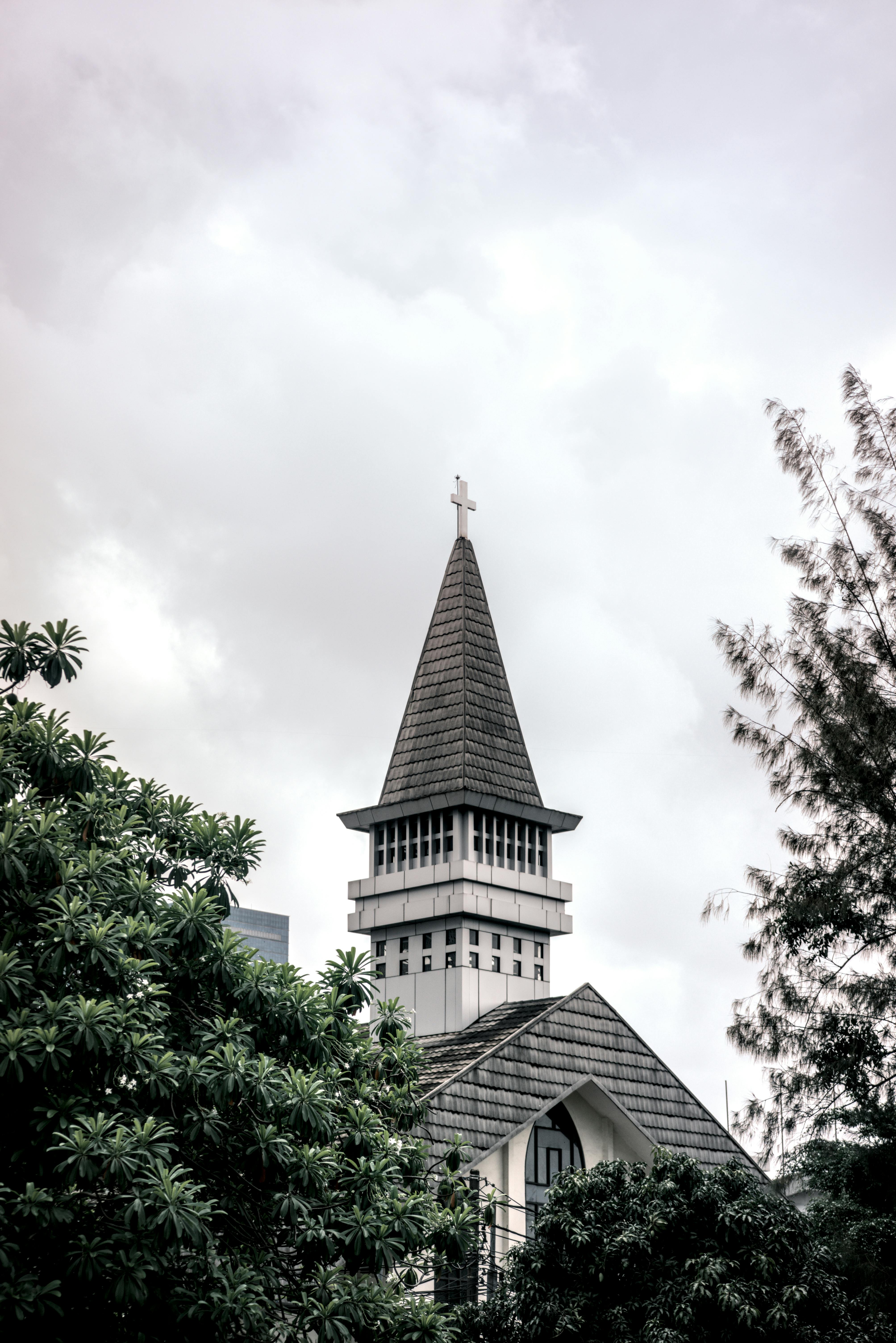 Ücretsiz Bulutlu bir gökyüzüne karşı haçlı Gotik tarzı bir kilise kulesi, yemyeşil bitki örtüsüyle çevrili. Açık havada, hüzünlü bir mimari simge. Stok Fotoğraflar