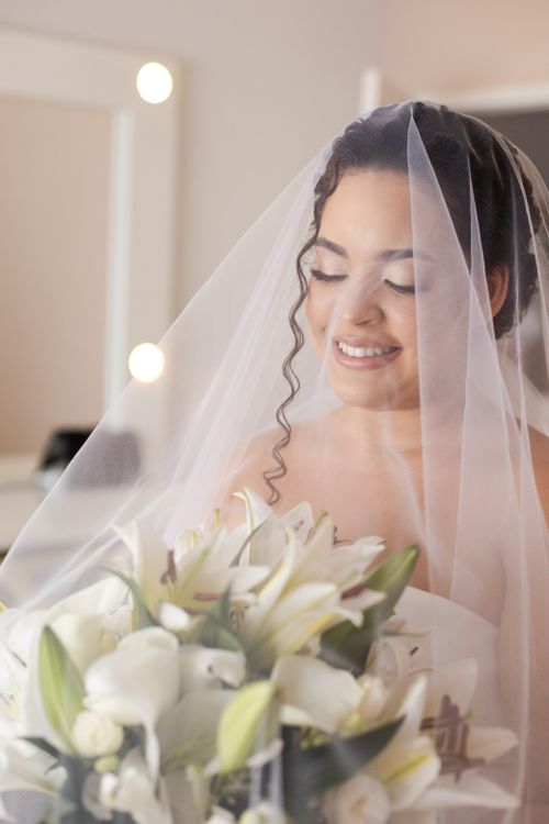 Free Bride in veil smiling while holding a bouquet of white lilies indoors. Stock Photo