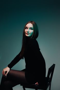 Stylish woman in black dress posing on chair in studio with moody lighting.