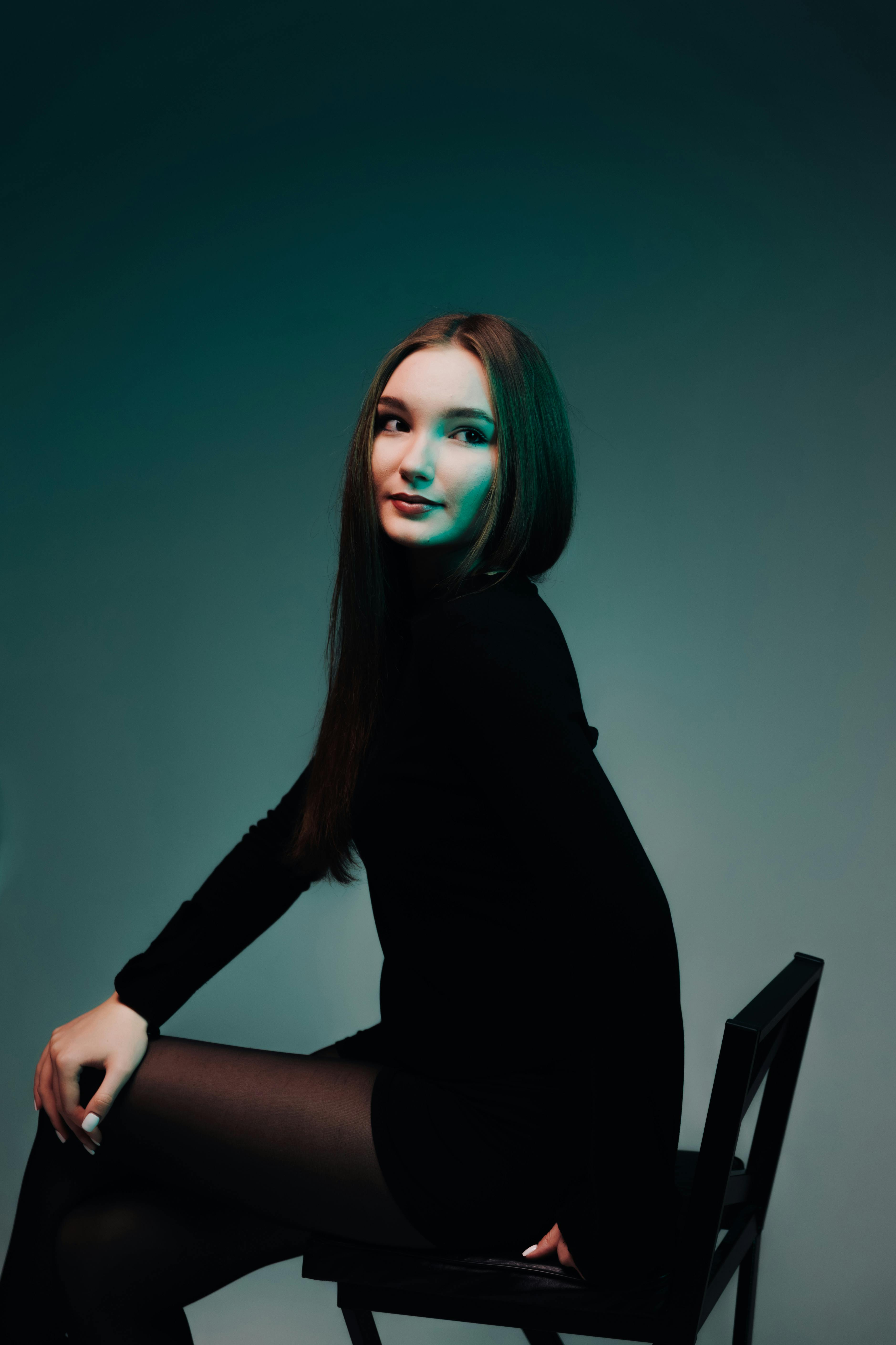 Gratis Stilfuld kvinde i sort kjole poserer på stol i studie med stemningsfuld belysning. Lagerfoto