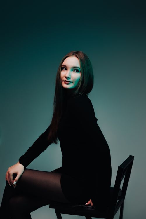 Free Stylish woman in black dress posing on chair in studio with moody lighting. Stock Photo