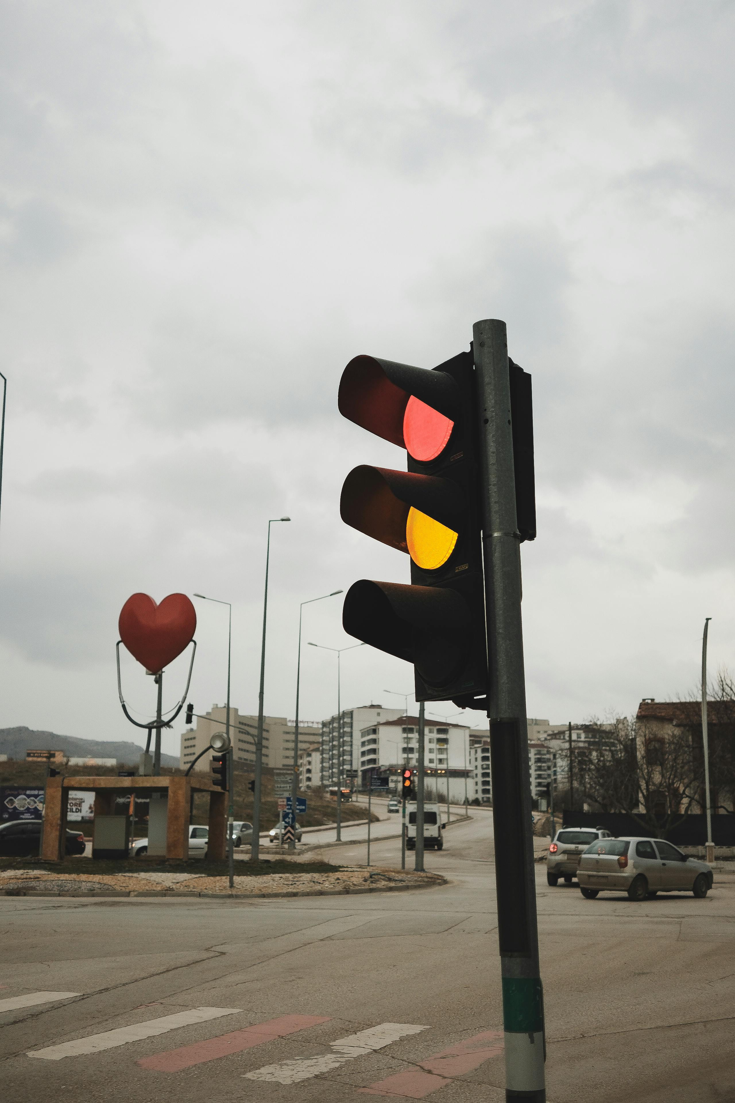 Gratis Trafiklys med røde og gule lys på en overskyet dag i en by med hjerteformet skulptur i baggrunden. Lagerfoto