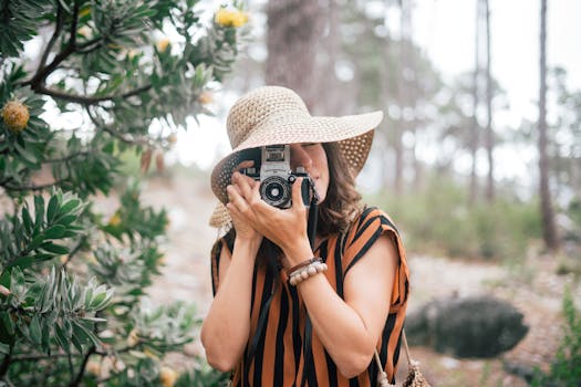 A woman photographer wearing a sun hat takes photos amidst lush greenery in an outdoor setting.