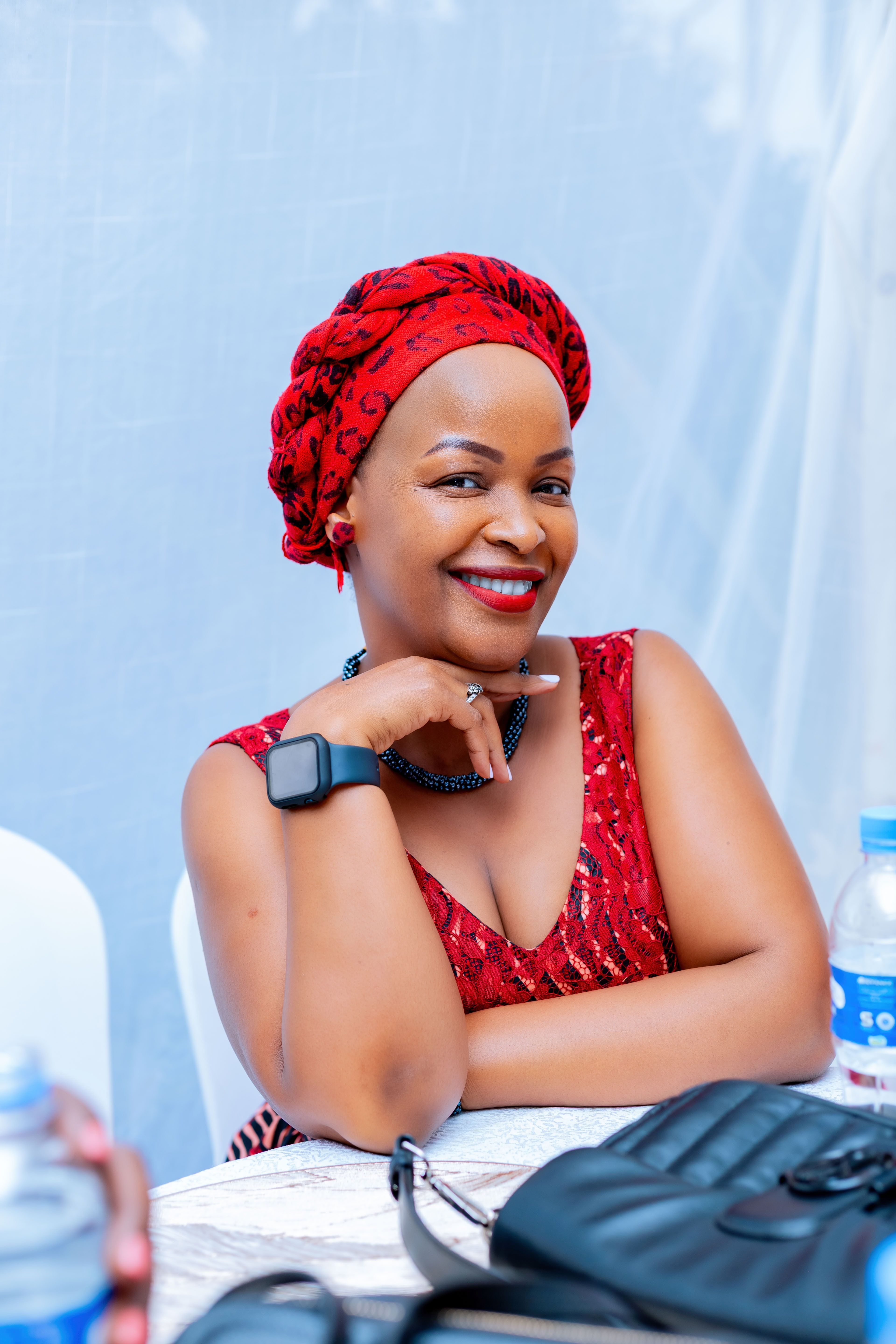 Smiling woman in a red dress and headscarf sitting at an indoor gathering with a table setup.