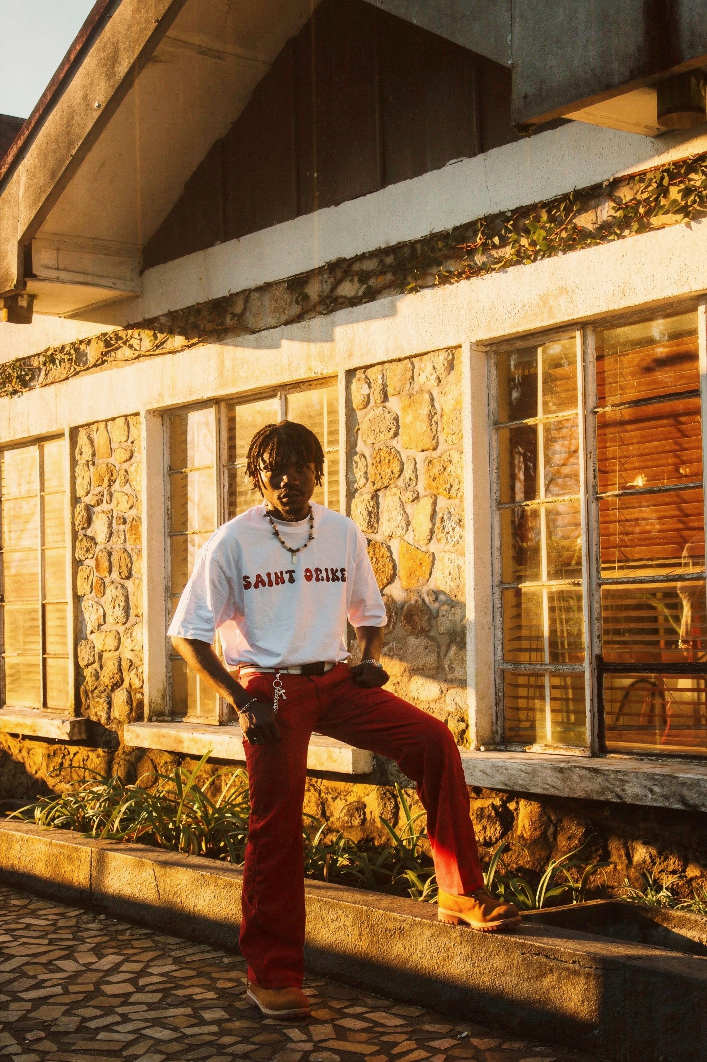 A man in red pants poses confidently by a stone wall in warm golden lighting.