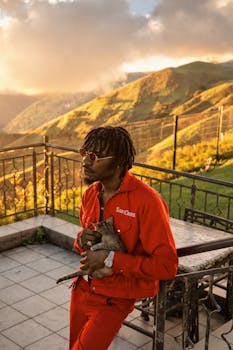 A man in red clothes and sunglasses holds a cat on a beautiful terrace with mountain views at sunset.