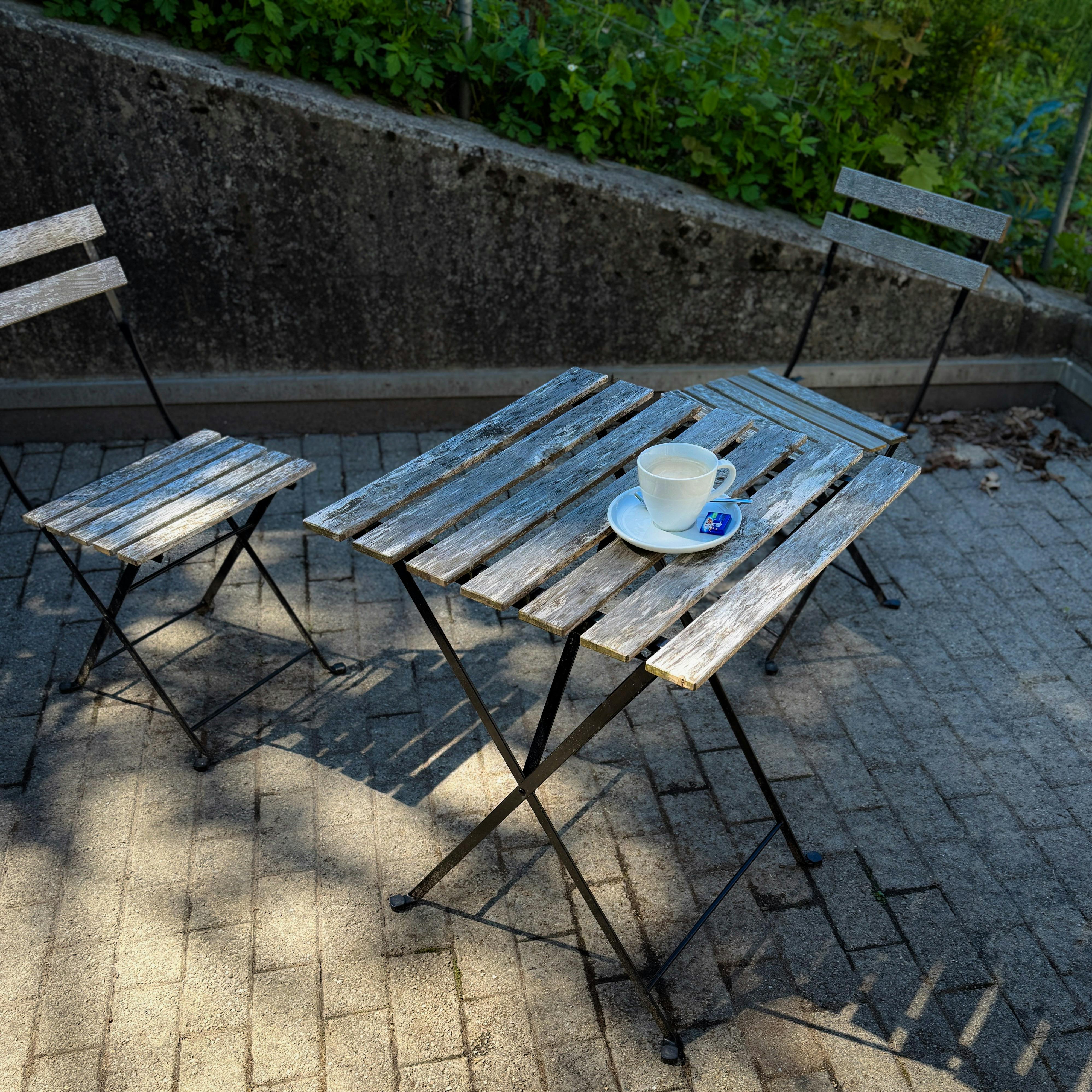 Gratis En sjarmerende utendørskafé med værbitte tremøbler og en kopp kaffe på en solrik terrasse. Arkivbilde