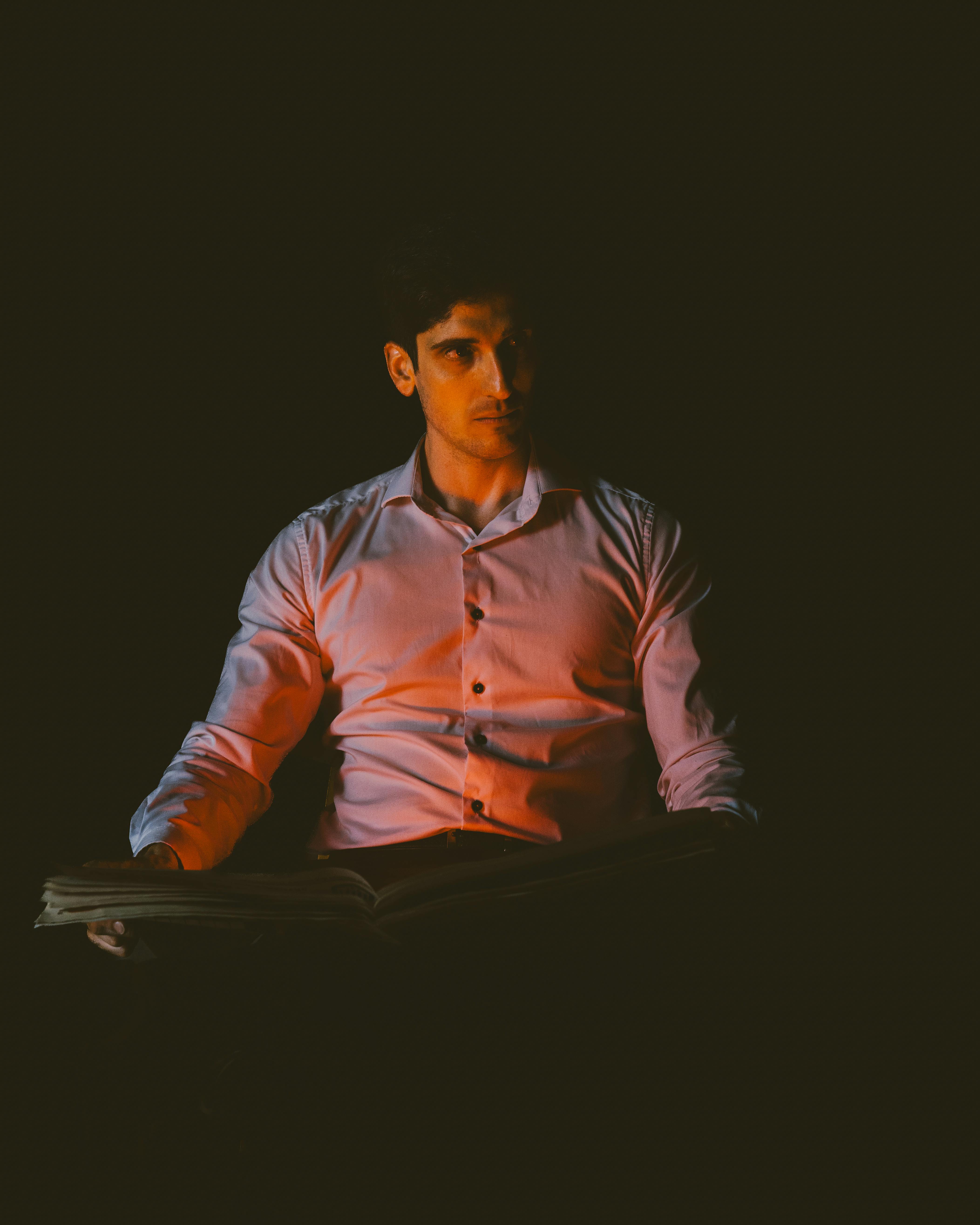 Bezpłatne Młody mężczyzna czyta w przyćmionym świetle, co wywołuje tajemniczy, kontemplacyjny nastrój. Zdjęcie z galerii