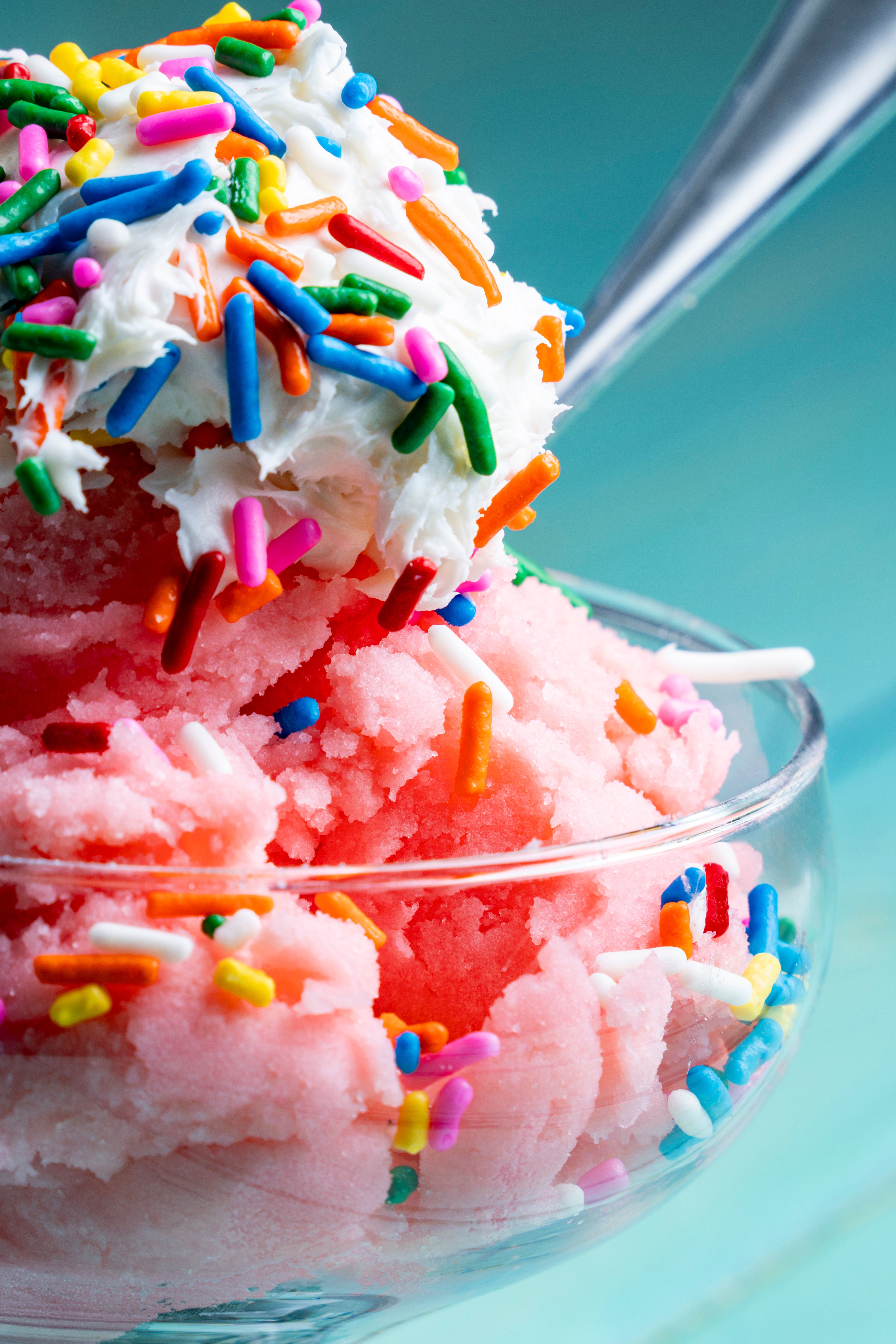 Close-up of a pink frozen dessert topped with whipped cream and colorful sprinkles in a glass bowl.