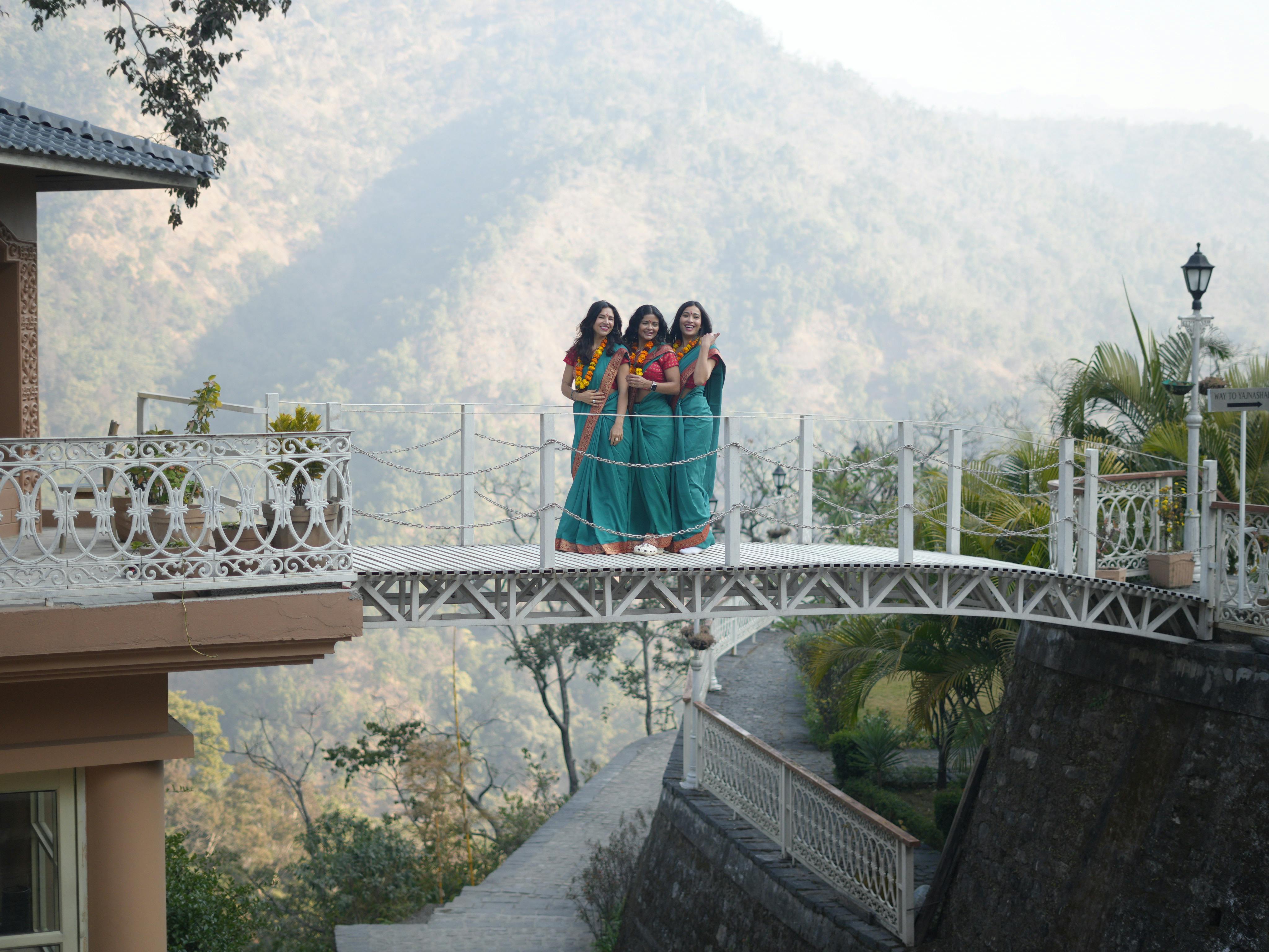 Three women in traditional dresses standing on a bridge in Rishikesh, India, with scenic mountain views.