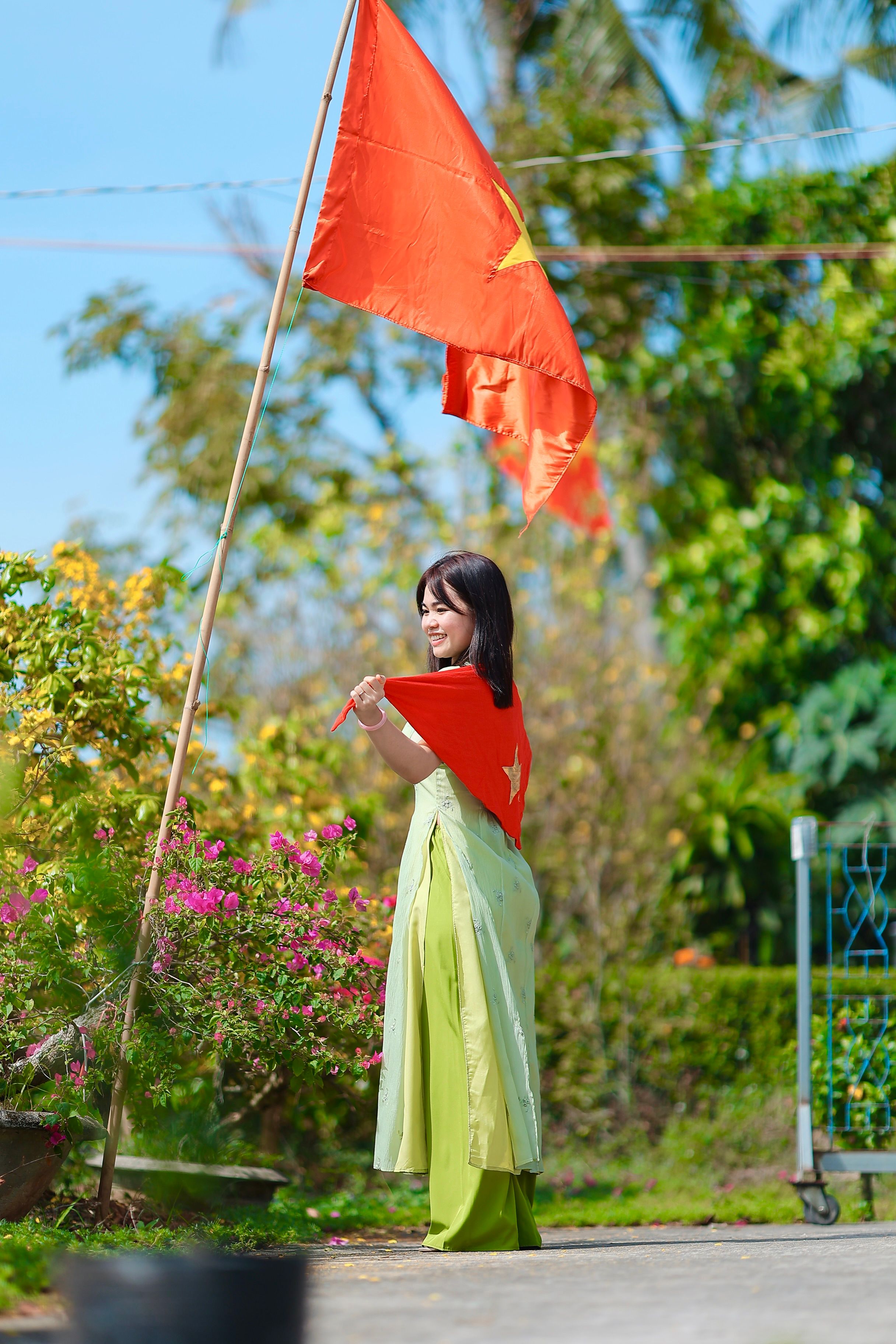 A young woman in traditional attire holds a Vietnamese flag in a vibrant garden setting.