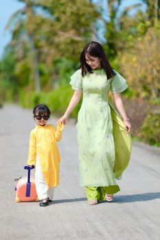 A woman and a child in traditional dresses walking outdoors with a suitcase.