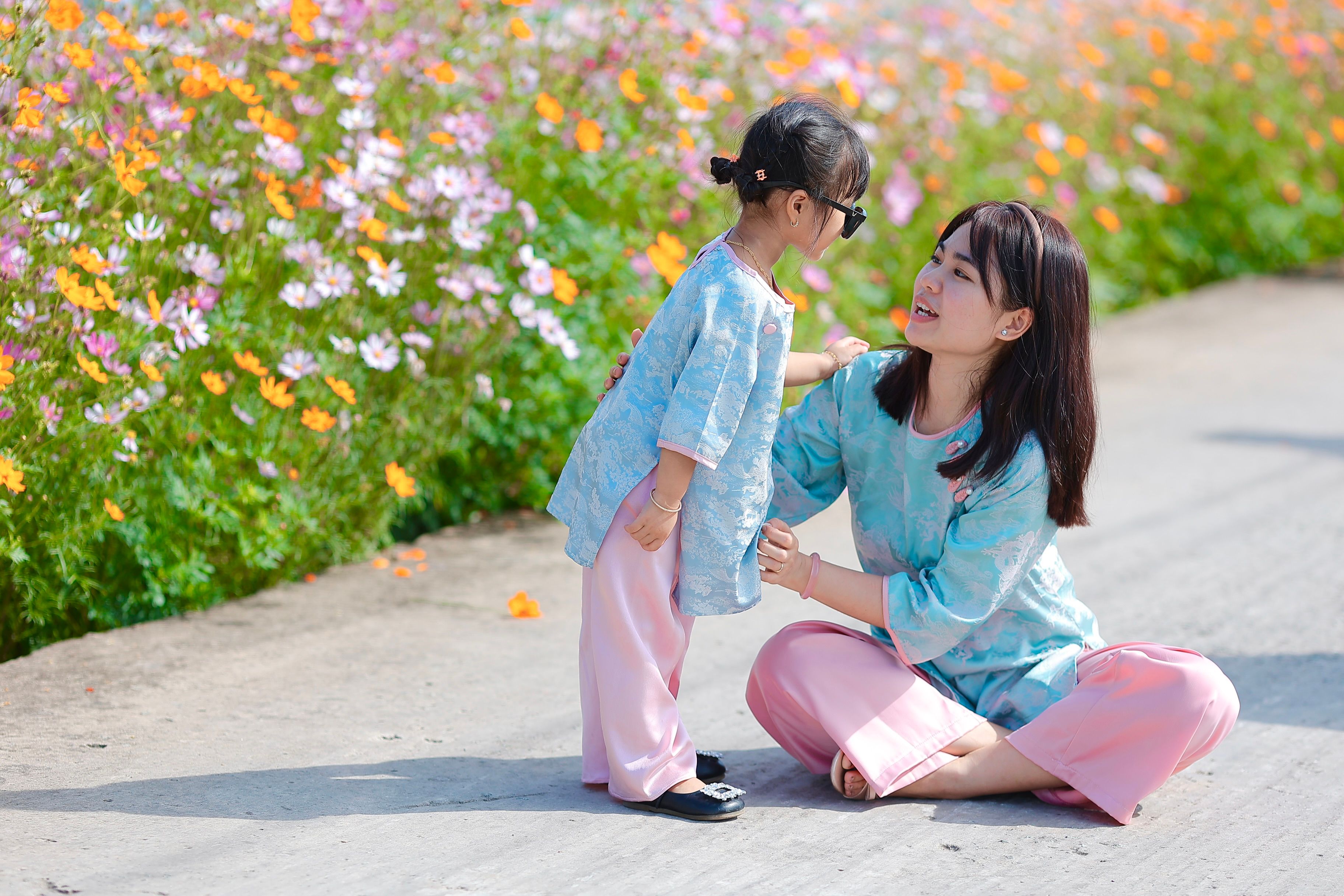 A mother and daughter in blue and pink outfits enjoying a sunny day outdoors with colorful flowers.