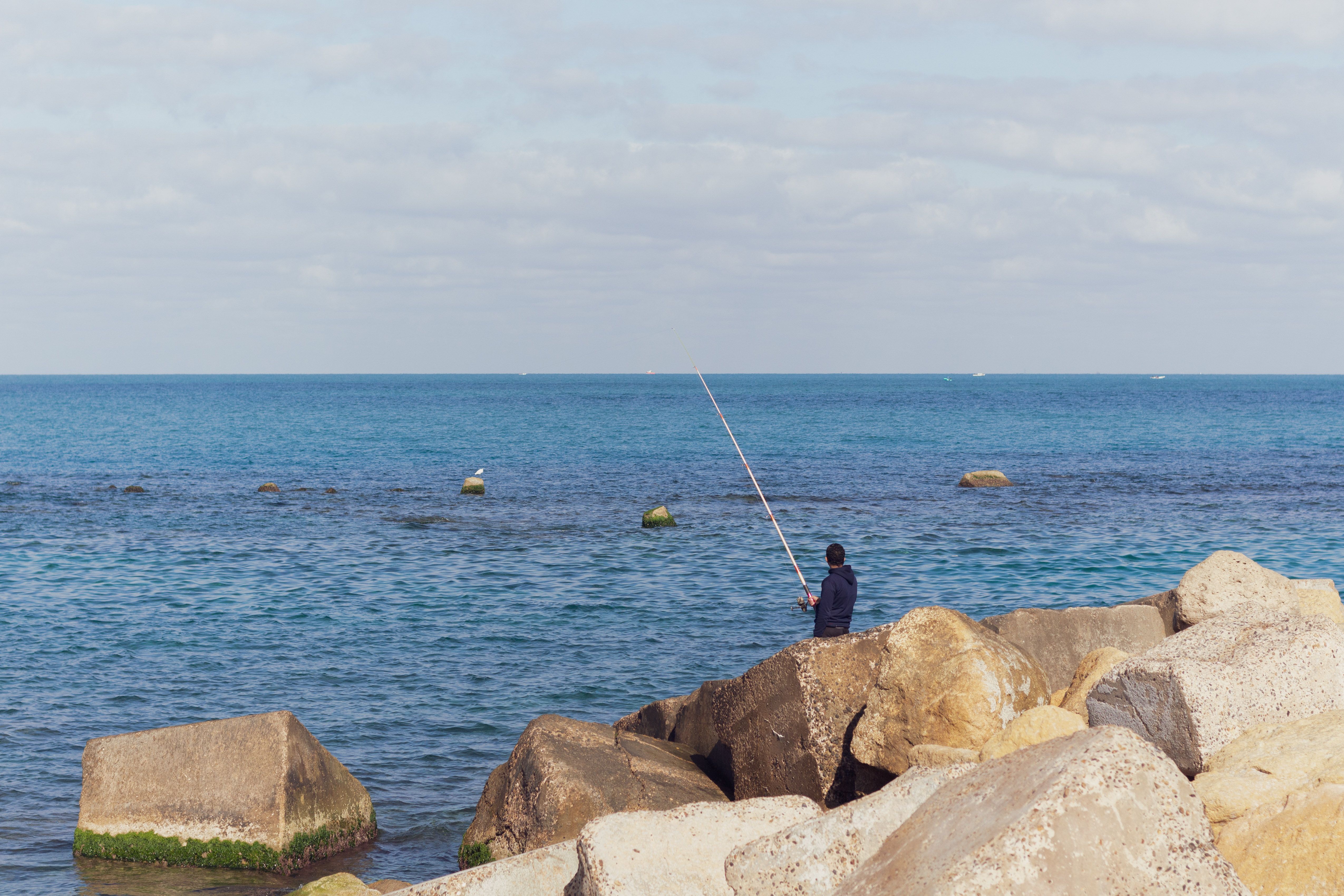 Gratis Un hombre solitario pesca en la costa rocosa de Alejandría, Egipto, bajo un cielo claro de verano. Foto de stock