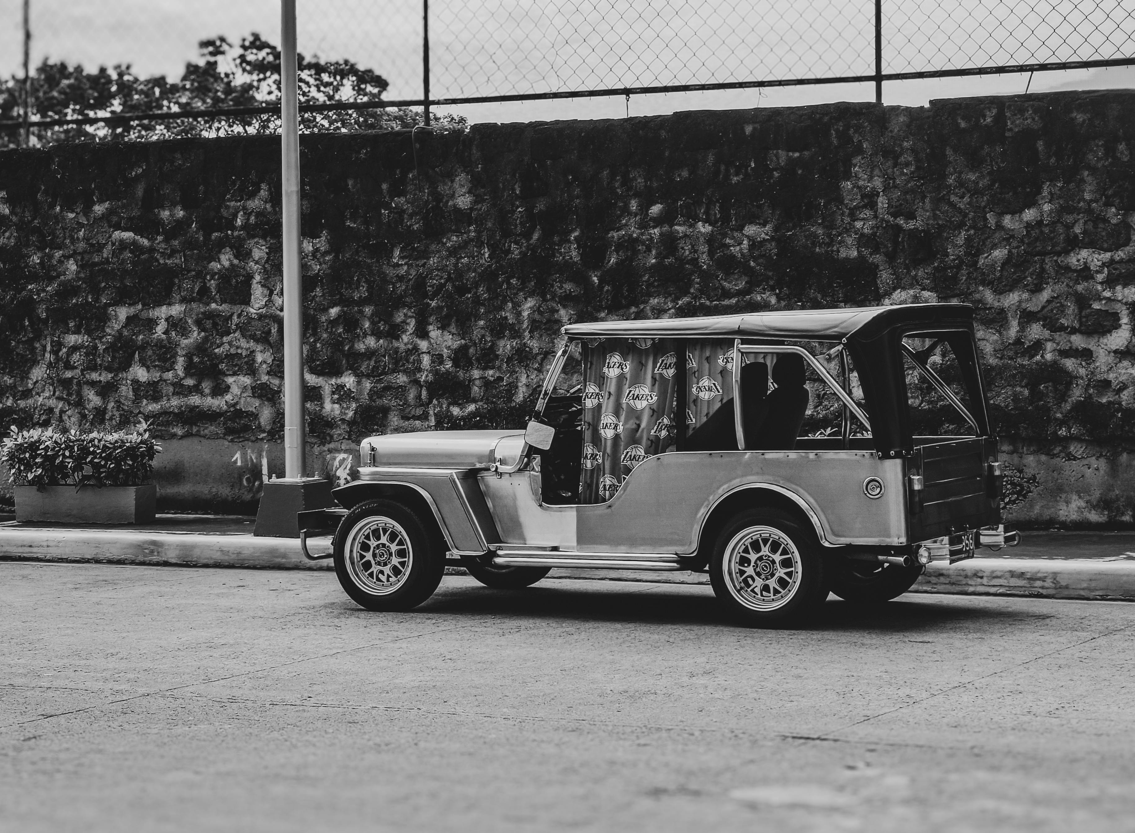 Gratis Sebuah Jeep klasik terparkir di jalan perkotaan yang tenang dengan latar belakang tembok batu dalam warna monokrom. Foto Stok