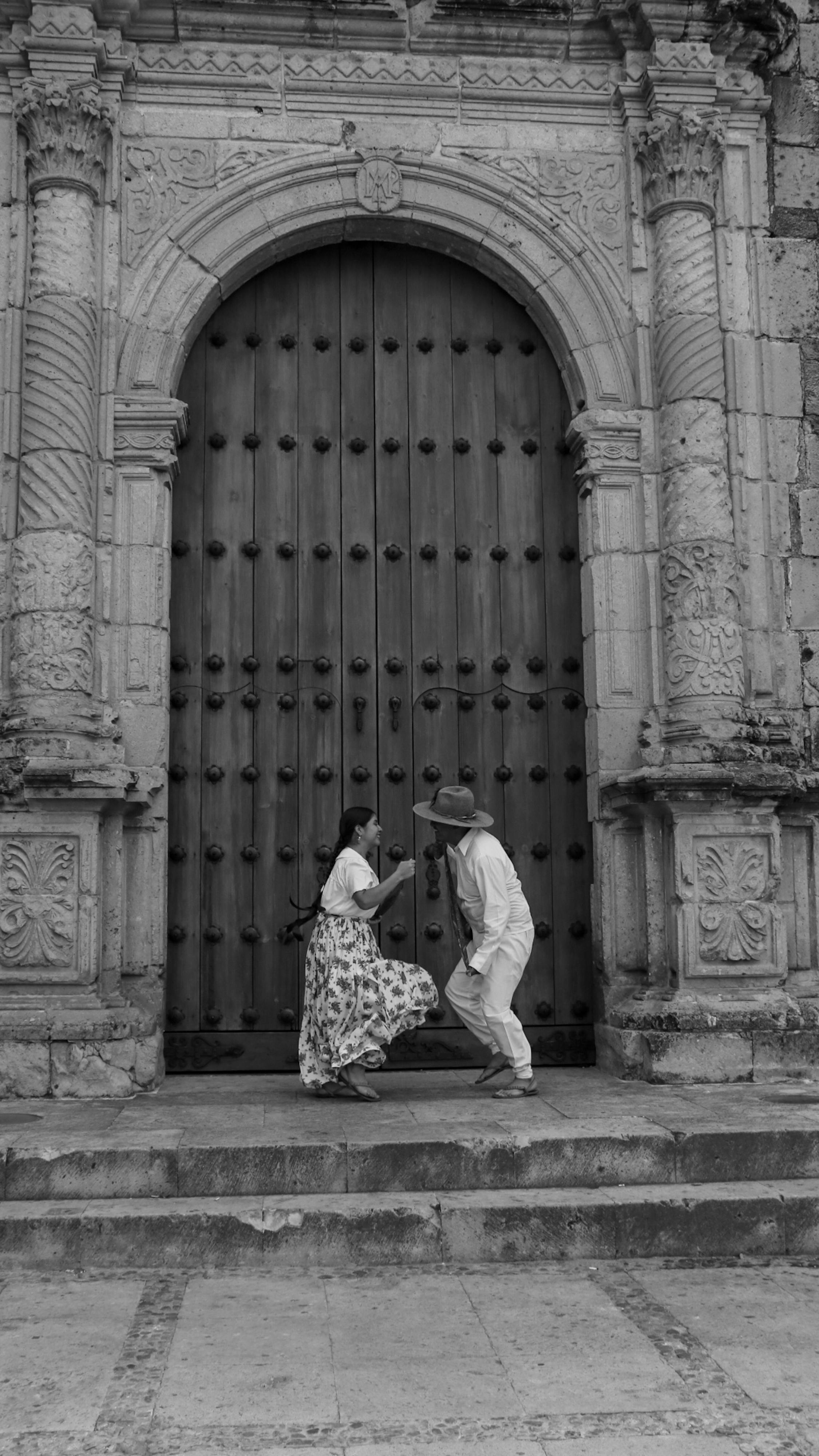 Gratis Gambar hitam putih sepasang kekasih yang sedang menari di depan sebuah gapura batu besar dengan ukiran yang rumit. Foto Stok