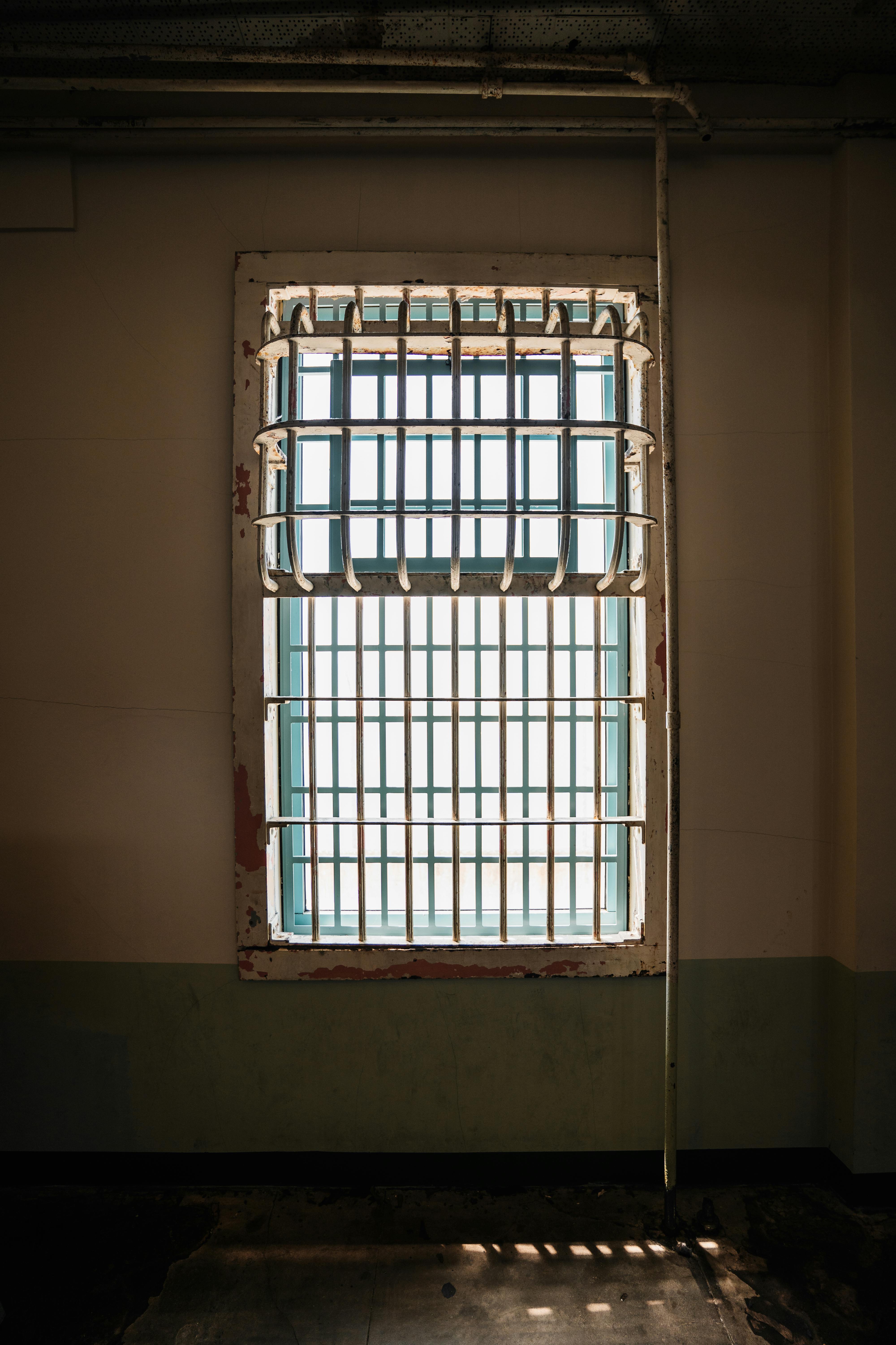 Kostenlos Ein vergittertes Fenster im Inneren einer historischen Gefängniszelle, durch das Sonnenlicht Schatten wirft. Stock-Foto
