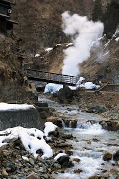 A picturesque hot spring with a wooden bridge over a rocky stream, surrounded by snow.