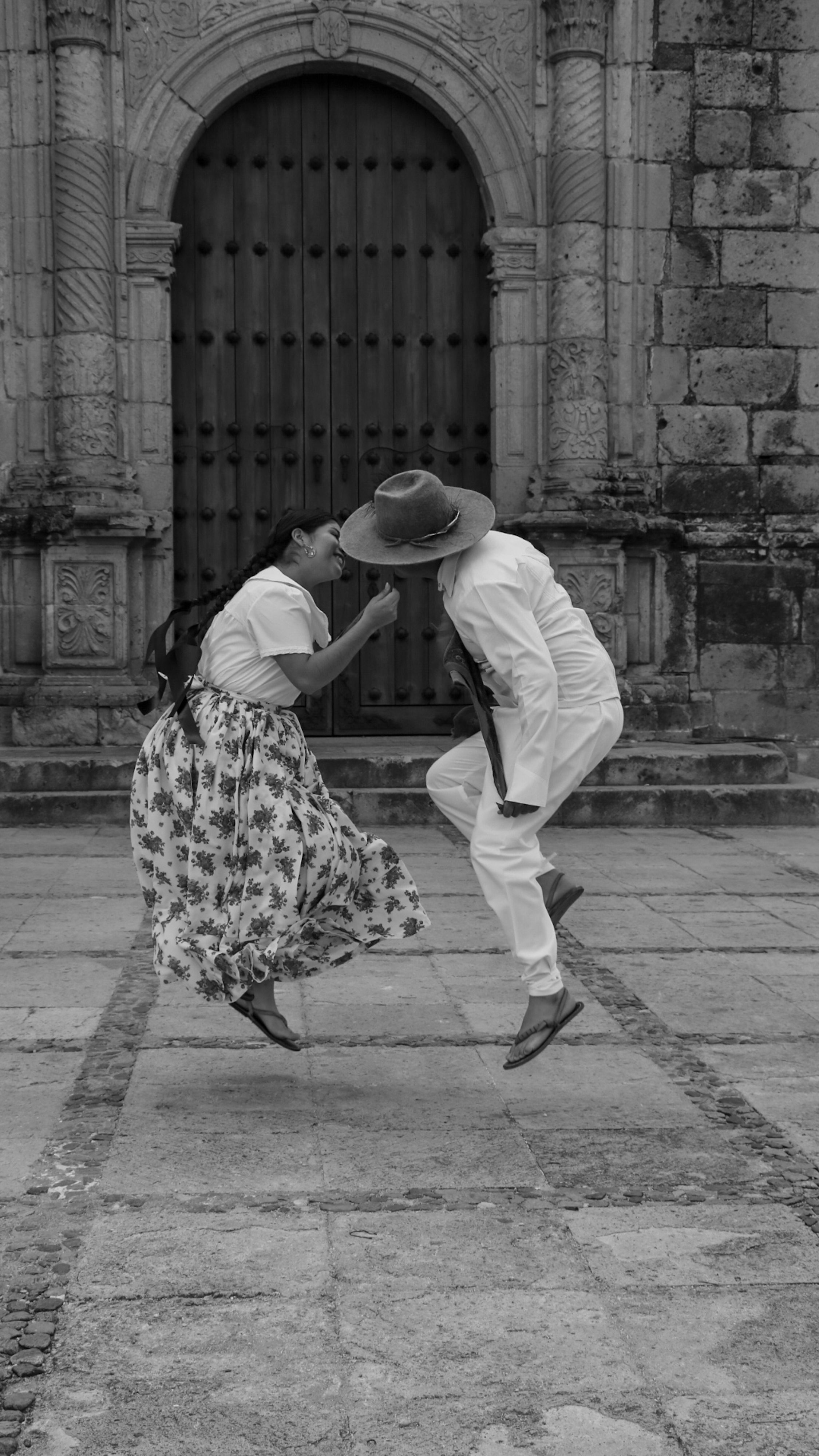 Black and white photo of a couple in traditional attire jumping near an ornate stone doorway.