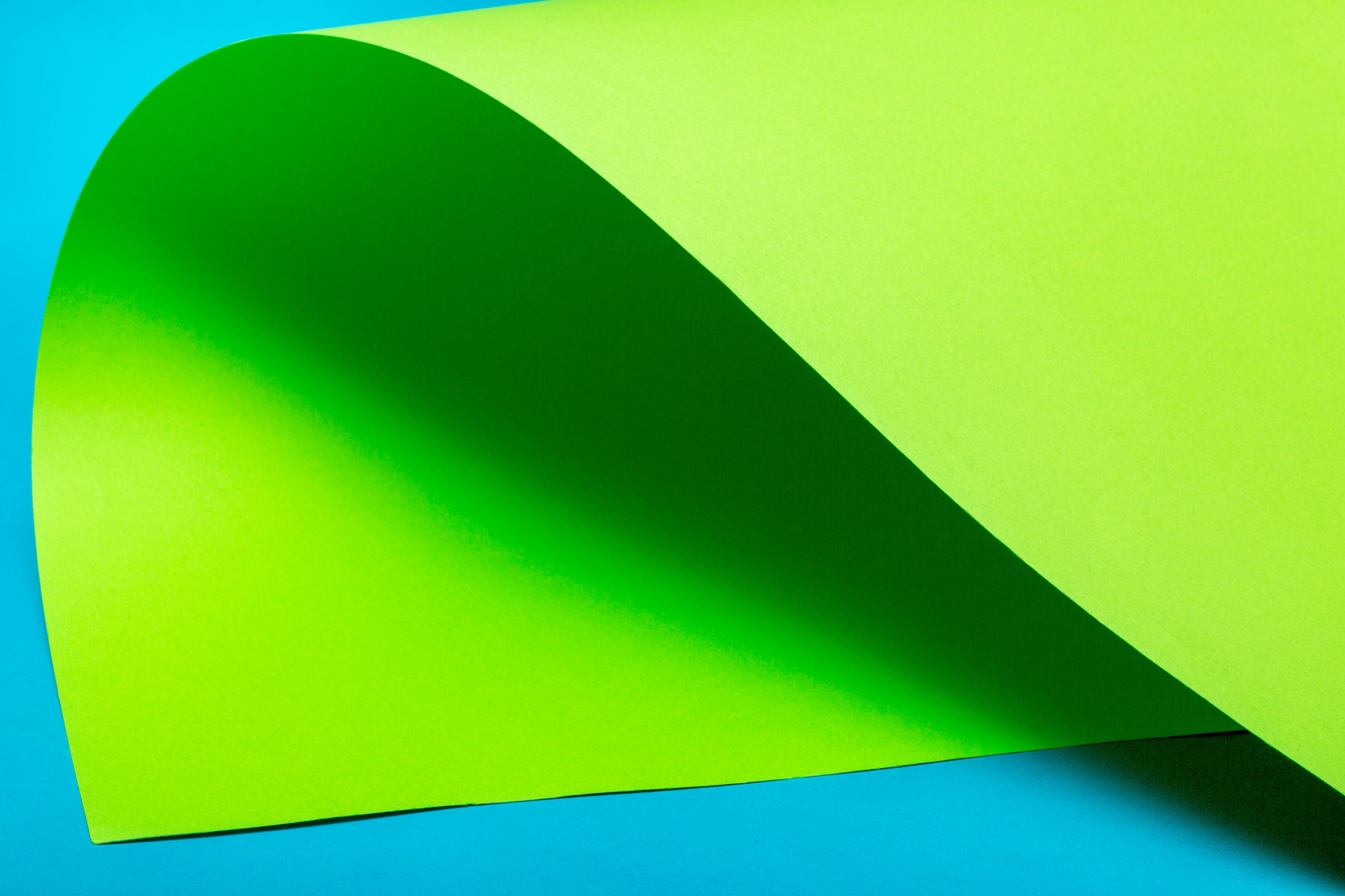 Gratis Et levende billede af et foldet, lyst grønt papir oven på en blå overflade, der skaber en abstrakt form. Lagerfoto