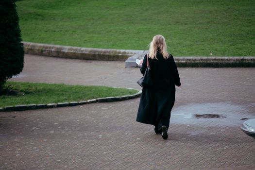 A woman in a black coat walks alone in a peaceful Dublin park on a rainy day.