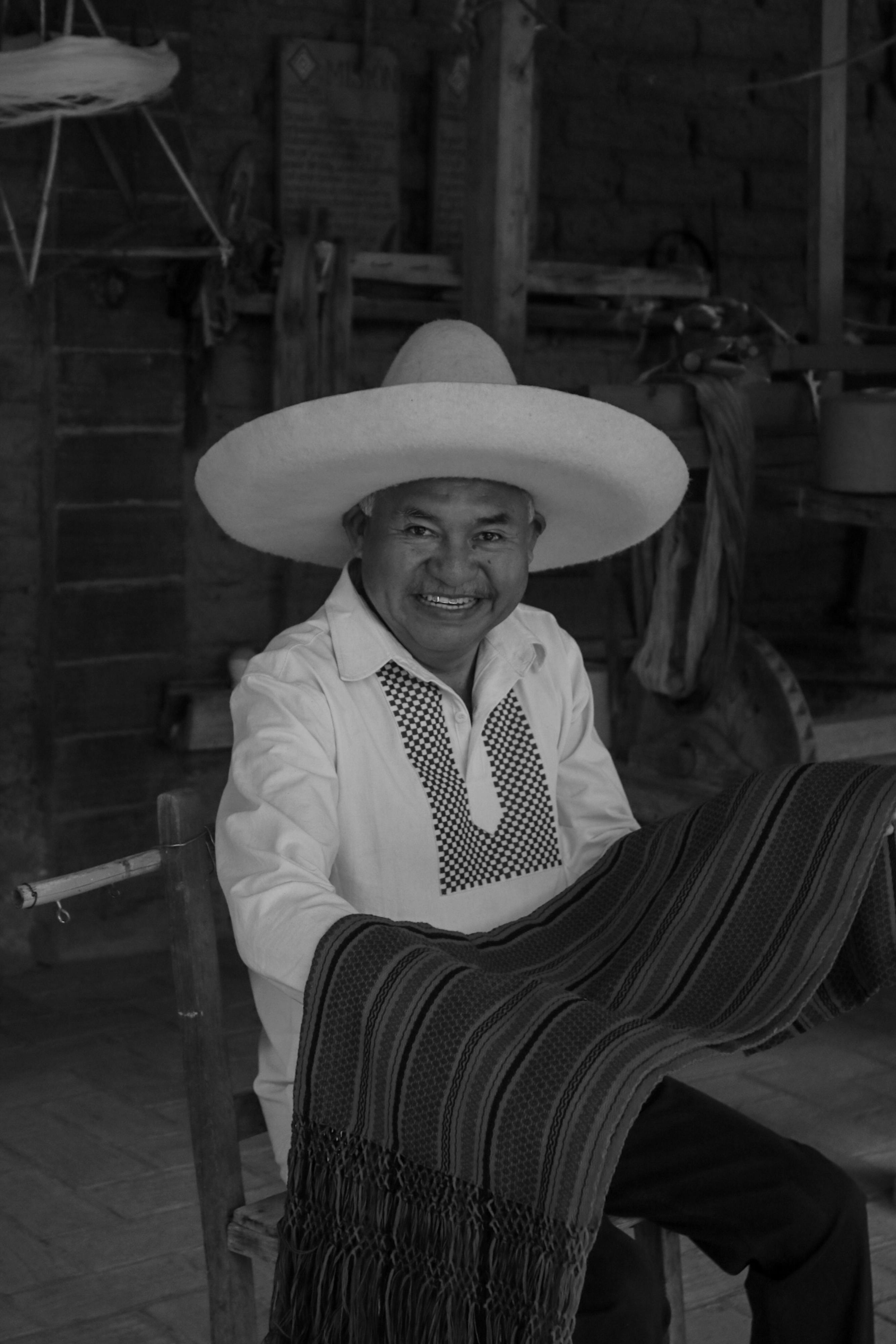 Bezpłatne Uśmiechnięty mężczyzna w tradycyjnym stroju zajmujący się tkaniem w pomieszczeniu, uchwycony na czarno-białym zdjęciu. Zdjęcie z galerii