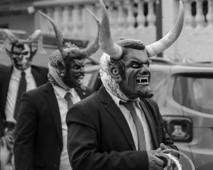 Black and white photo of a parade with people wearing devil masks and suits, depicting a cultural festival.