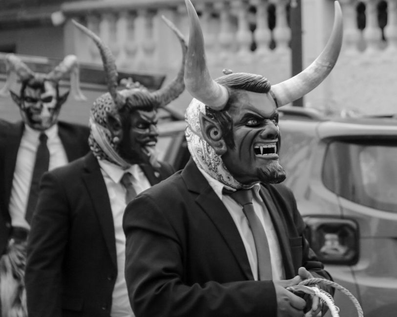 Black and white photo of a parade with people wearing devil masks and suits, depicting a cultural festival.