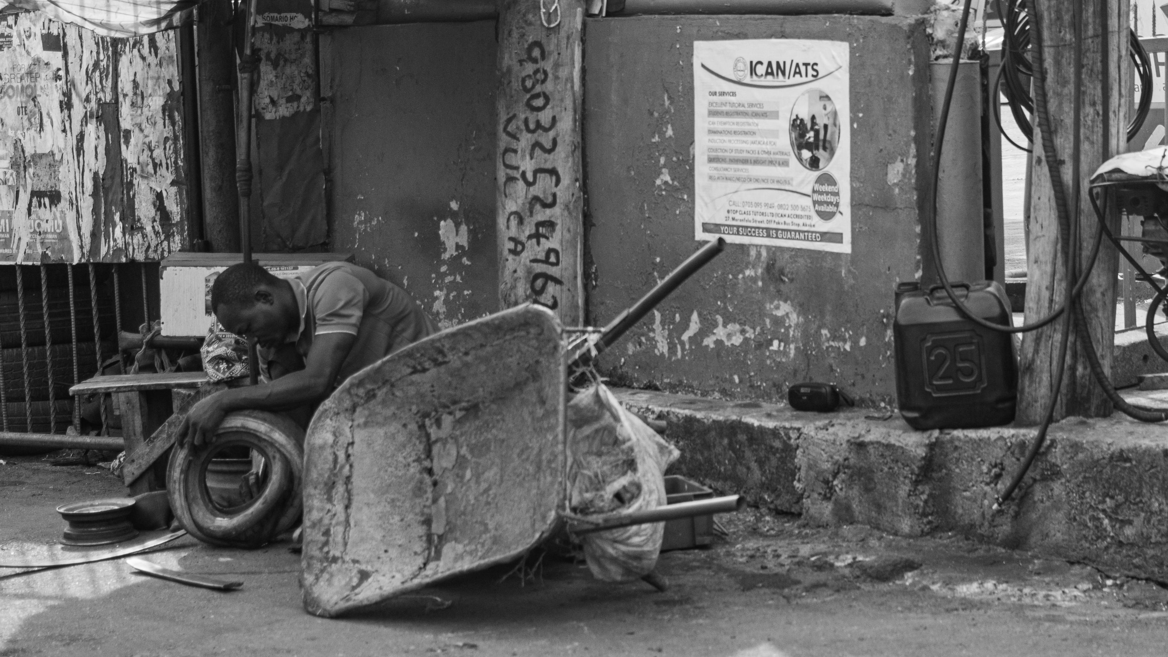 Gratuit O fotografie alb-negru sinceră a unei persoane care repară o anvelopă la colțul unei străzi, evocând viața urbană de zi cu zi. Fotografie de stoc