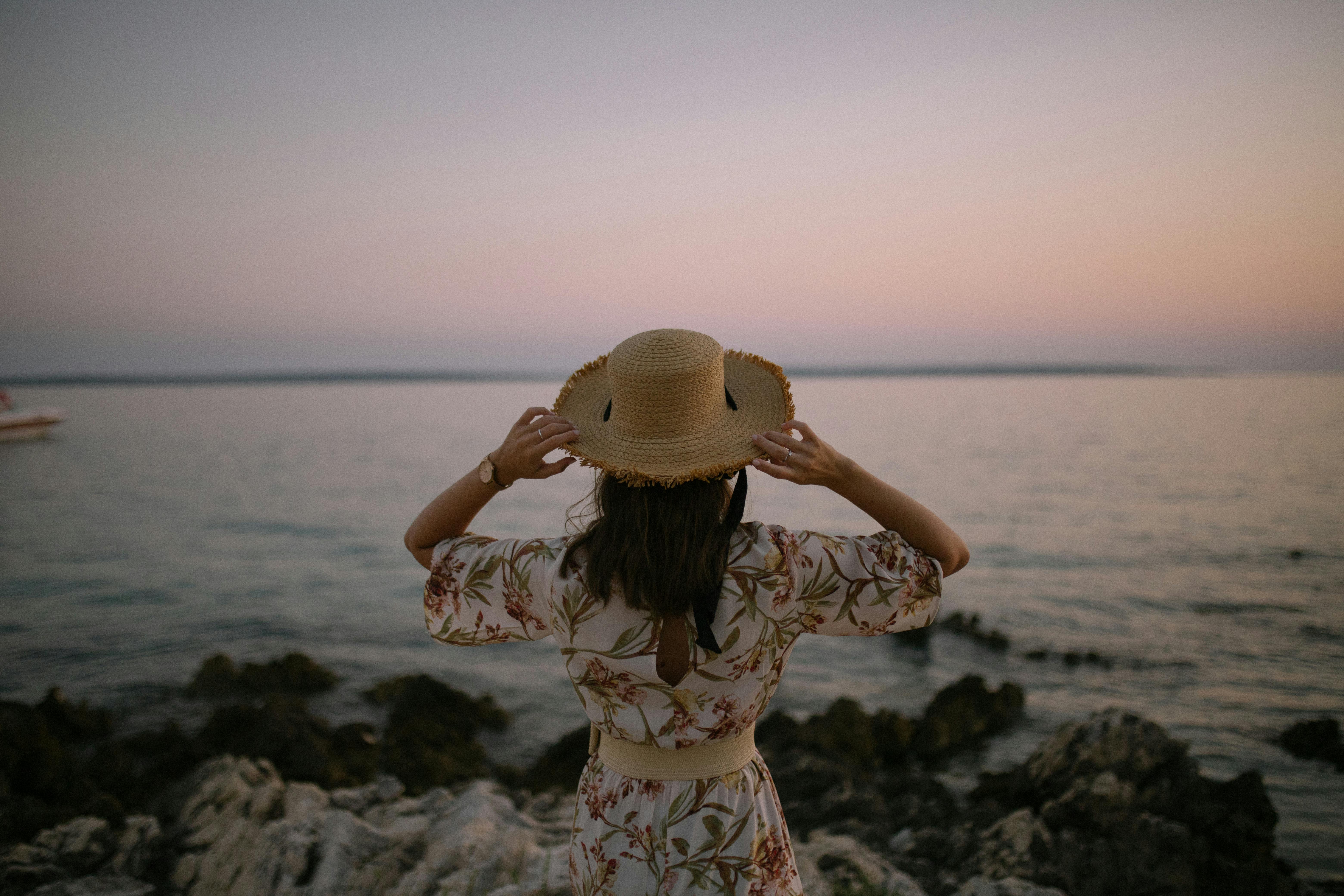 Ücretsiz Çiçekli bir elbise ve hasır şapka giymiş bir kadın, gün batımında sakin bir sahil manzarasının tadını çıkarıyor. Stok Fotoğraflar