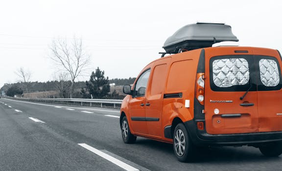 Bright orange van traveling on an empty highway with a roof box on a cloudy day.