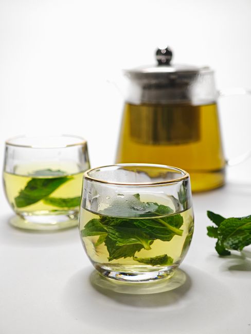 A glass teacup of pale green mint tea steeping with fresh mint leaves and a cinnamon stick on a wooden tray