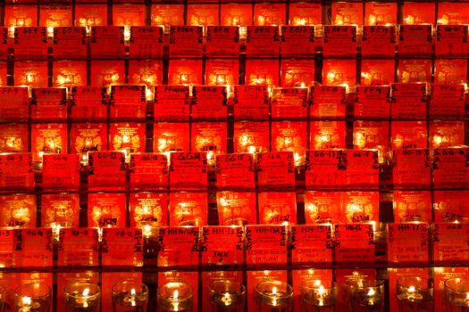 A collection of votive candles set against a wall of red cards, creating a warm and spiritual ambiance.