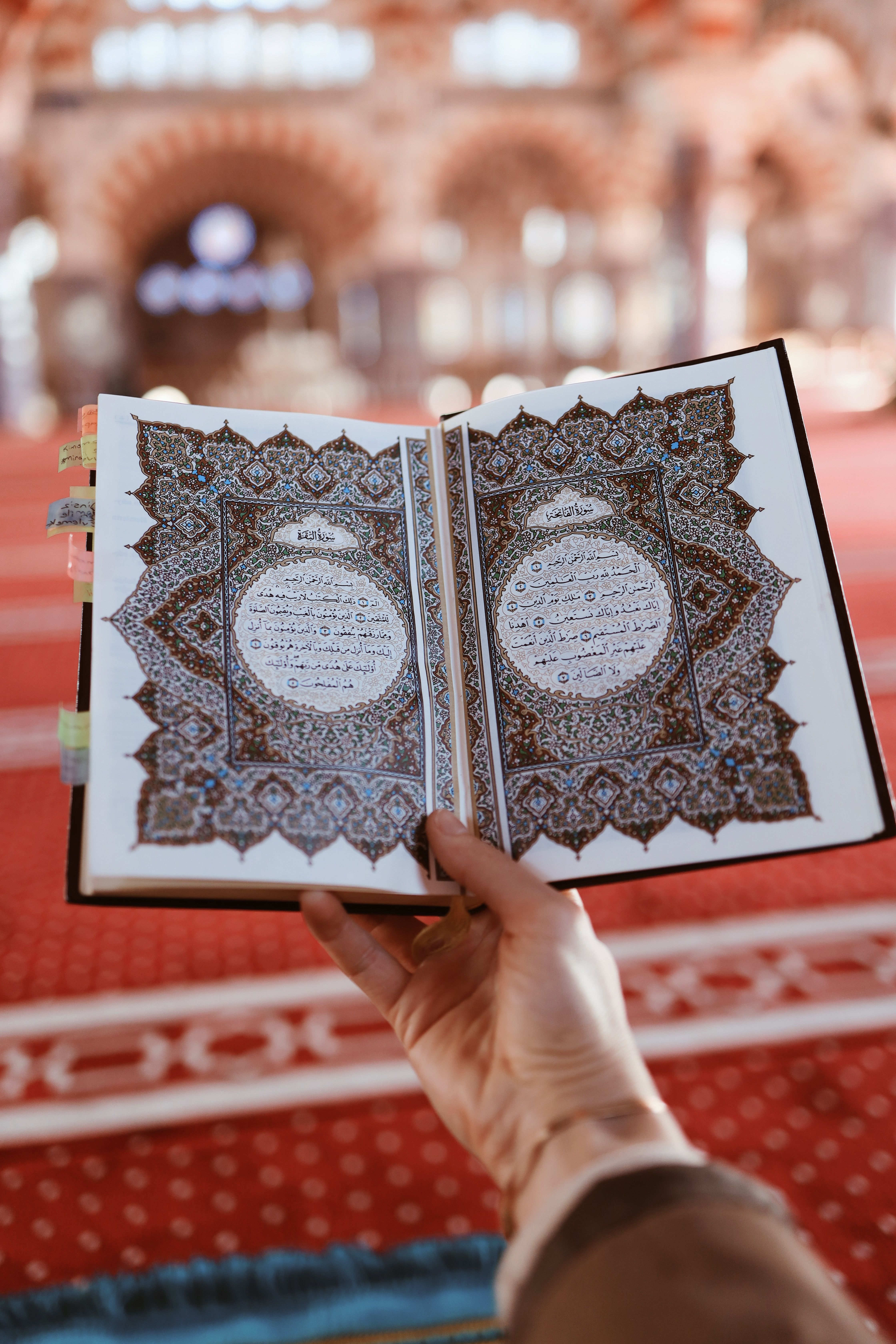 A hand holds an open Quran inside an ornate mosque with arched ceilings.