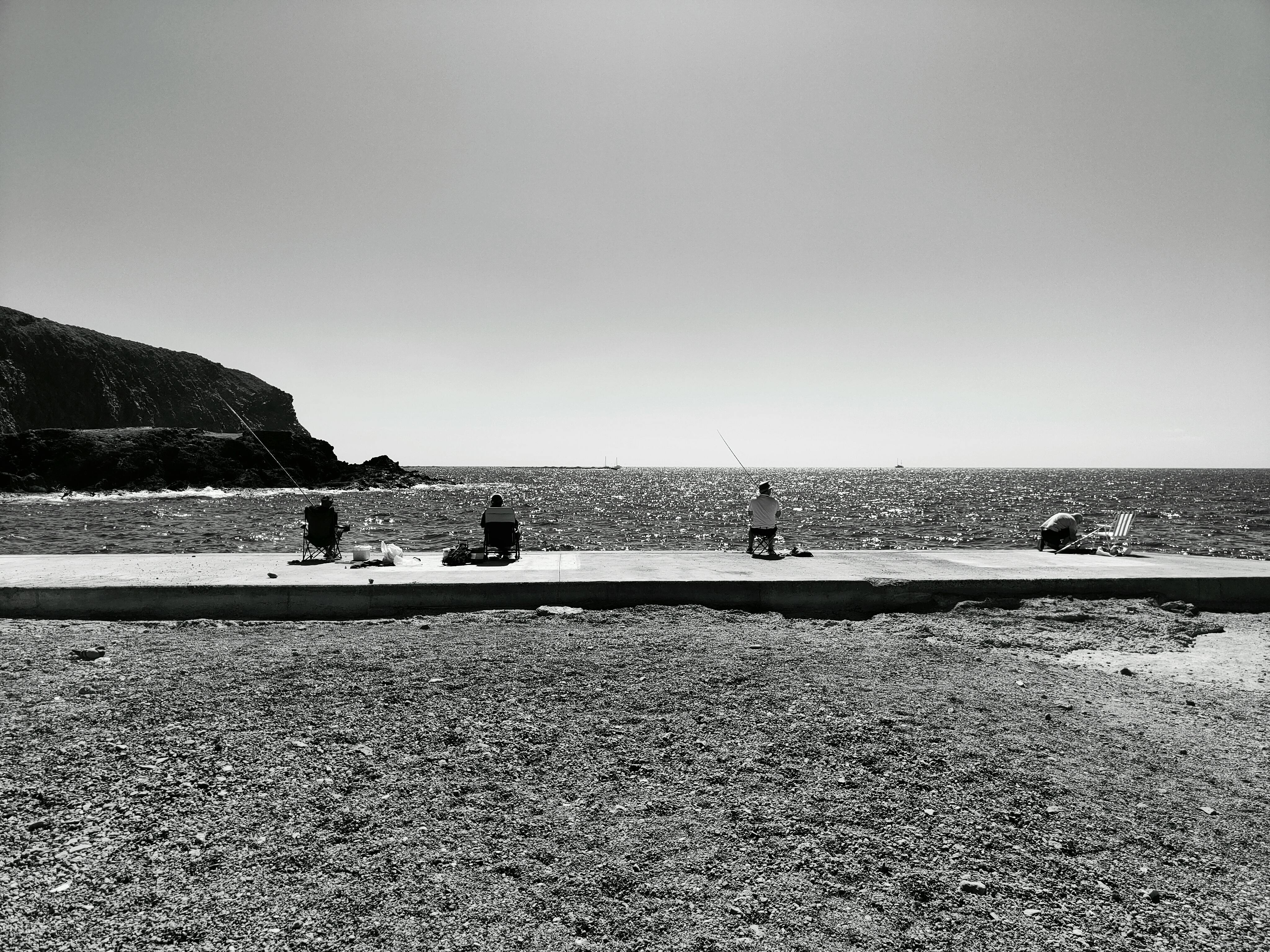 Gratis Tre personer fisker ved havet under en klar himmel, fanget i monokrom. Lagerfoto