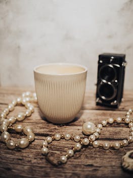 A vintage camera and pearls adorn a wooden surface beside a ceramic cup, conveying elegance.