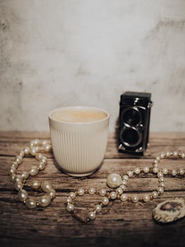 Elegant still life featuring a ceramic cup, pearls, and vintage camera on wooden surface.