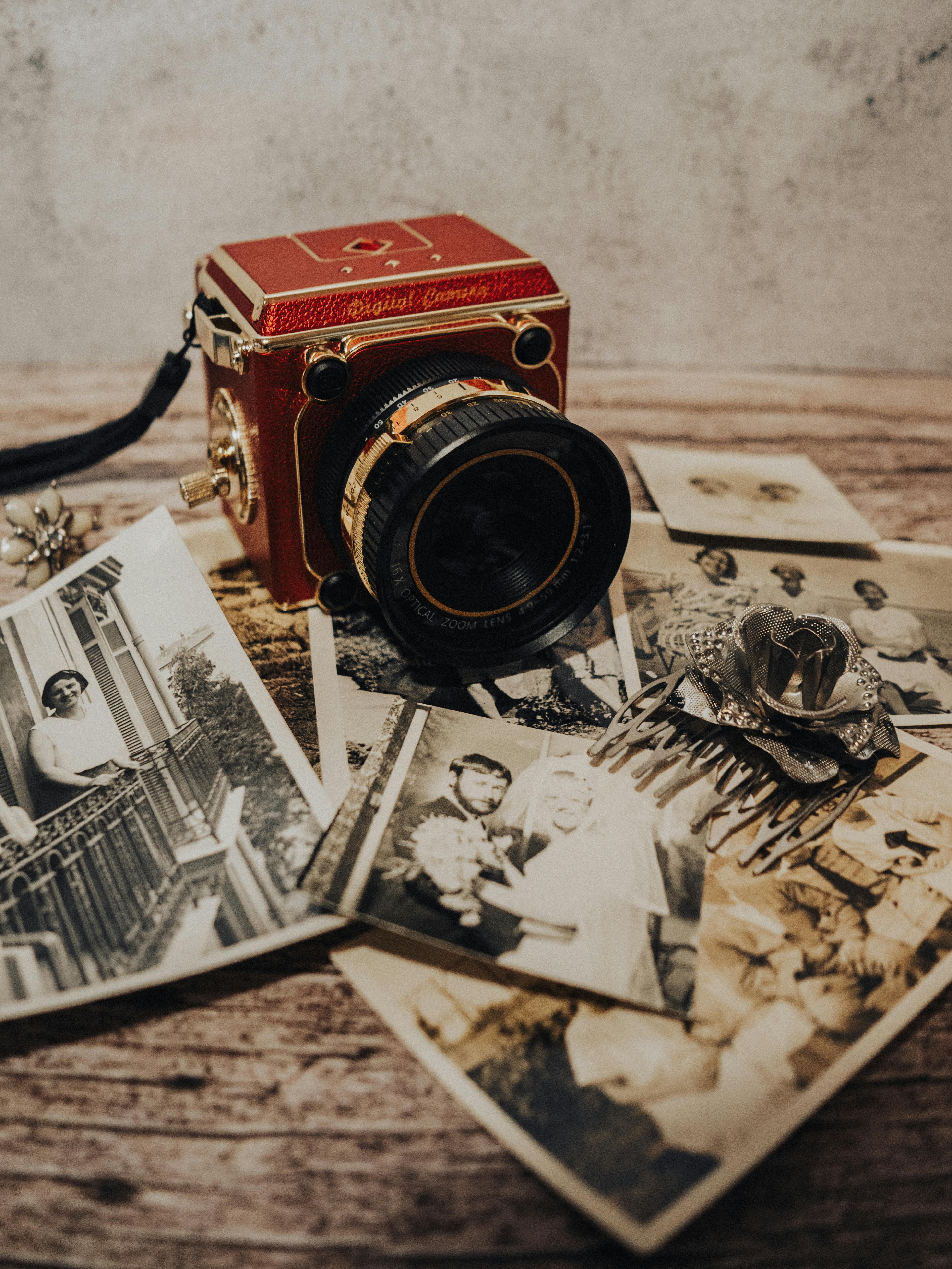 Bezpłatne Czerwony, zabytkowy aparat fotograficzny otoczony starymi czarno-białymi zdjęciami na drewnianym stole. Zdjęcie z galerii