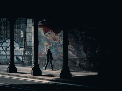 A person walks through a shadowy urban underpass with colorful graffiti walls.